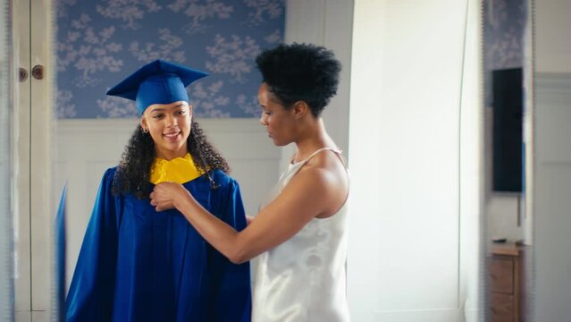 Proud mother celebrating with teenage daughter wearing graduation robes reflected in mirror at home 