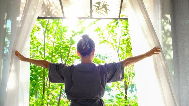 The woman was at the window in the bedroom. She opened the curtains on the window. In the morning and she looks at the view of mountains and trees at sunrise.