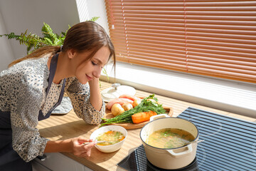 Young woman eating chicken soup in kitchen - obrazy, fototapety, plakaty