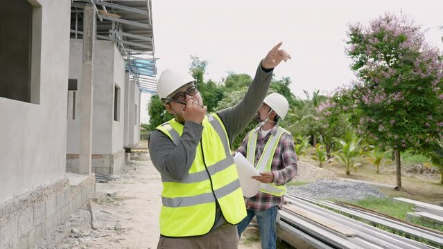 Asian construction supervisor Use a wireless radio Talk to high crane operator. to control crane Man standing using walkie-talkie at home construction site Wear helmet and reflective safety equipment.
