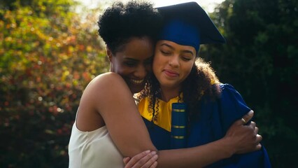 Proud mother celebrating with teenage daughter wearing graduation robes outdoors - shot in slow motion