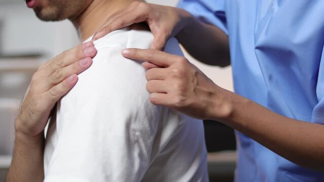 Doctor diagnosing man's shoulder pain in hospital examination room. Nurse doing massage and physiotherapy to treat patient's shoulder.