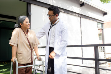 Young doctor assistance elderly patient while guidance therapy with walker in garden at hospital, caregiver or physician support senior and practicing walking with walker, medical and rehabilitation.