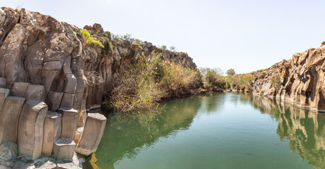 A small  lake with fish framed by hexagonal rocks with overgrown bushes and trees on the banks in Yehudia National Natural Park in northern Israel - Powered by Adobe