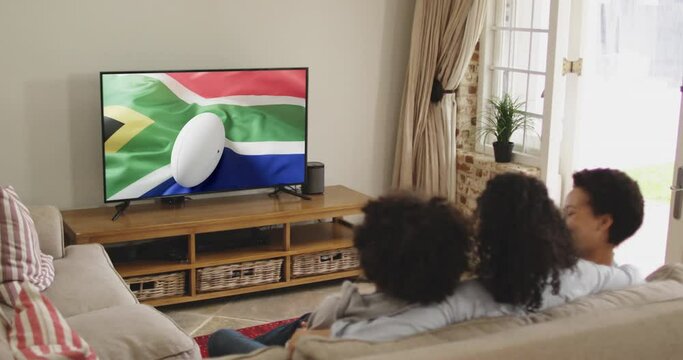 Biracial family watching tv with rugby ball on flag of south africa on screen