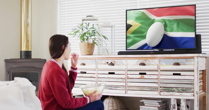 Caucasian woman watching tv with rugby ball on flag of south africa on screen