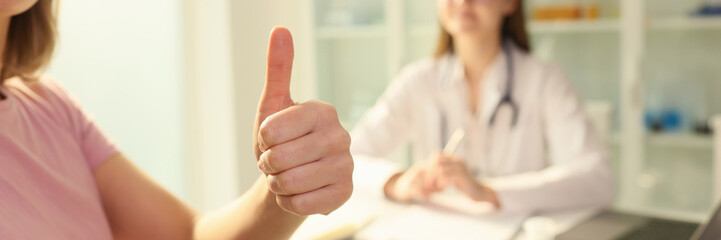 Woman patient shows thumbs up while sitting near female doctor in clinic.