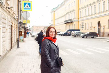 smiling young caucasian female woman in winter clothes in red scarf on street of European historical city, crosswalk. three quarter length portrait. tourism