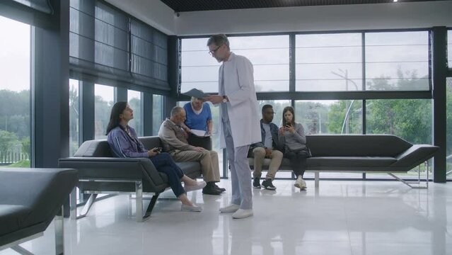 Diverse multicultural people sit on couches in clinic waiting area, wait for doctors appointment. Doctor speaks with patients about medical test results. Medical staff works in modern medical center.