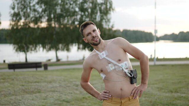 Topless man with heart monitoring sensors does warm-up in park at camera, arc shot. Slender guy with holter monitor checks heart function under stress. Person bends and jerks with his arms.