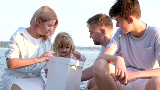 Happy family celebrating little girl's birthday on the seashore mom brings a box with cake girl opens a gift dad mom brother and sister on the ocean shore. on a sunny day