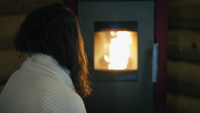 A young Caucasian woman is sitting by an electric pellet fireplace wrapped in a blanket.