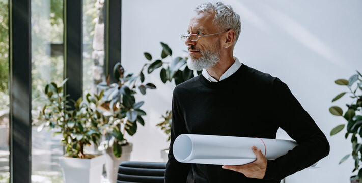 Confident mature architect carrying blueprint while standing in the creative office