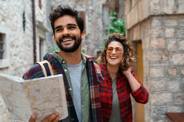 Beautiful young couple walking with a map in the city. Two friends spending vacation by exploring the streets of Mediterranean old town. Smiling man holding map and guiding his girlfriend.
