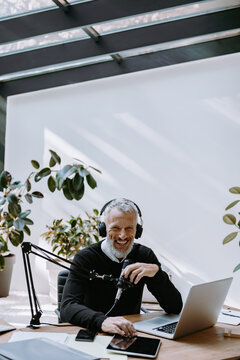 Joyful mature podcaster in headphones using microphone while broadcasting from studio