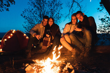 Two female and two male friends on camping trip, sitting close to the fire in a forest and enjoying their time together.