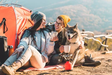 Foto op Plexiglas Photo of a happy young couple and their dog camping in the mountains on a beautiful autumn day and drinking a cup of tea outdoors. © Dorde