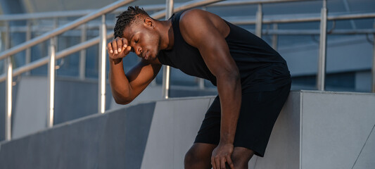Exhausted African man in sportswear leaning hands on knees while resting after training outdoors