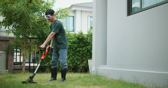 Asian man mows the grass in the backyard with an electric lawn mower. Gardener mowing lawn at backyard with brush cutter. Male cutting grass with an electric scythe. Lawn mowing