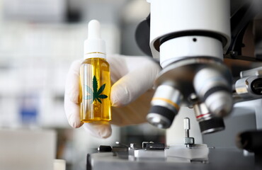 Focus on male hand in gloves holding glass bottle with cbd. Laboratory interior with microscope. Medicine and medical marijuana concept. Blurred background