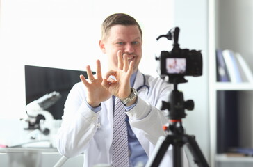 Portrait of smart doctor practitioner sitting in hospital office and performing approving gesture with hands to high-tech camera for vlog. Healthcare concept