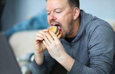 Portrait of male sitting indoors and eating big tasty hamburger full of mischievous calories and...