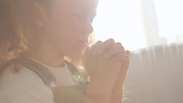 Close up of cute little girl sitting on cozy sofa at home keep hands in prayer hope believe ask good luck, hopeful pretty small child believer pray do religious superstitious gesture, faith concept