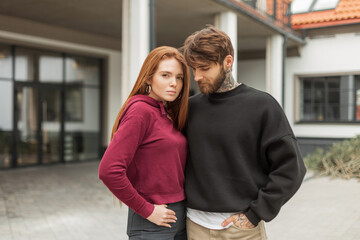 Fashionable beautiful stylish young couple in stylish clothes with a hoodie on the street. Handsome hipster man with beard and pretty red-haired girl stands outdoors