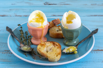 Soft boiled eggs in cups served with roasted bread, silver spoons, fresh thyme on blue plate.