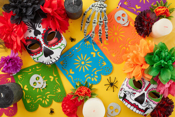 Joyful Day of the Dead. Top view flat lay of traditional masks with elaborate decorations, colorful garland, candles, cobwebs, spiders, skeleton hands on yellow background
