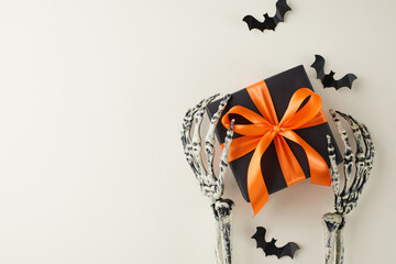 Bringing joy to Halloween with gifts for loved ones. Top view composition of black paper present box, spooky skeleton hands, scary bats on light grey background with commercial placeholder