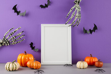 It's time to get set for Halloween. Side view of scary skeleton hands, pumpkins, spooky spiders and bats on the table on purple background with frame for text or advert