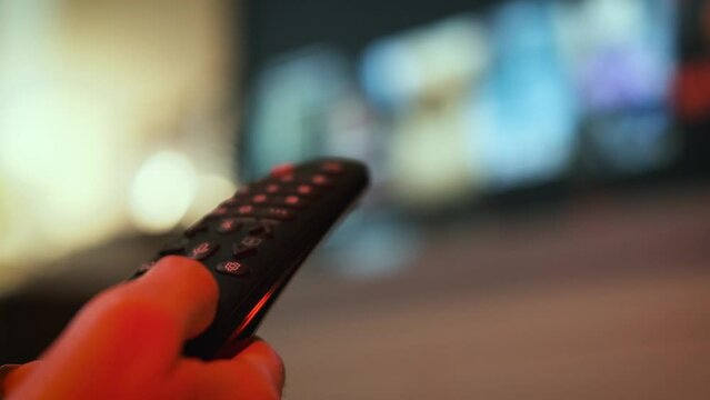 Woman hand selects internet tv channels with remote control. Close up view. Blurry tv at the background