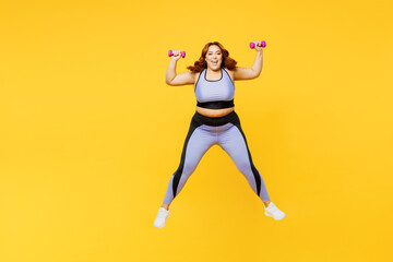 Full body excited young chubby plus size big fat fit woman wear blue top warm up train hold dumbbells jump high look camera isolated on plain yellow background studio home gym. Workout sport concept.