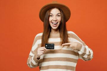 Full body young happy woman she wear striped sweater hat casual clothes hold in hand point finger on mock up of credit bank card isolated on plain orange red color wall background. Lifestyle concept.