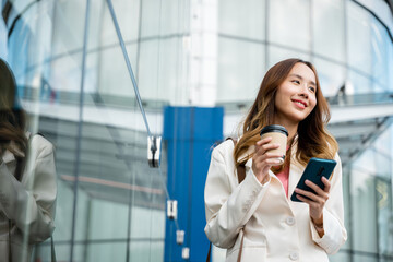 Young business woman smiling holding mobile phone with coffee take away going to work early in morning, Asian businesswoman with smartphone and cup coffee standing against street building near office