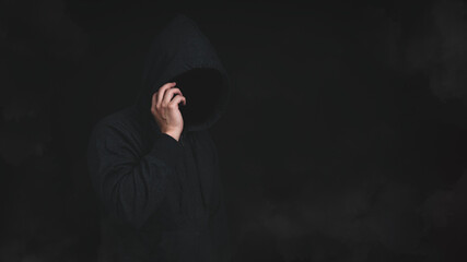 An unknown man in hood jacket using cellphone in dark background. Hacker, scammer or criminal using...
