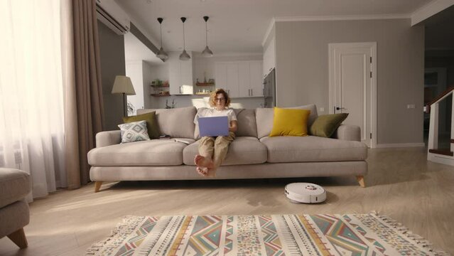 Woman working with laptop on sofa while robot vacuum cleaner cleans apartment. Cleaning robot performing daily vacuuming while pretty lady in sitting at sofa Automatic machine dusting off large spaces