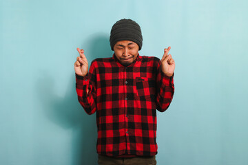 Hopeful young Asian man makes a finger crossed gesture, closes his eyes, isolated on blue background - obrazy, fototapety, plakaty