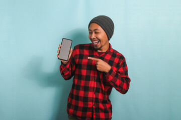 An excited young Asian man with a beanie hat and a red plaid flannel shirt points his finger at a mobile phone with a blank white screen, providing copy space, while standing against a blue background
