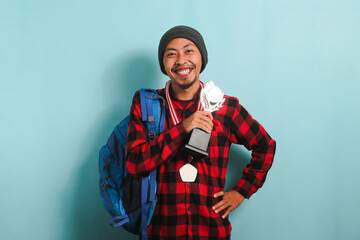 Happy young Asian man student showing a medal and trophy, isolated on a blue background