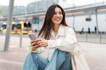 Waiting for the bus and for love. Bright cheerful young woman of Turkish mixed race in white casual clothes with a charming smile holding a phone in the street at public transportation stop - obrazy, fototapety, plakaty