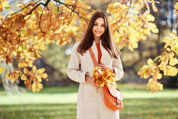 Autumn portrait of a beautiful happy smiling woman with yellow maple leaves in a white coat in the park