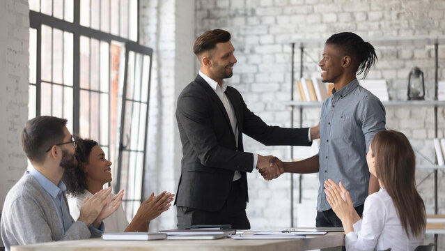 Smiling Caucasian businessman shake hand congratulate with good work achievement result african American male employee, boss handshake greeting with promotion excited biracial worker at meeting