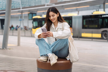 Waiting for the bus and for love. Bright cheerful young woman of Turkish mixed race in white casual clothes with a charming smile holding a phone in the street at public transportation stop