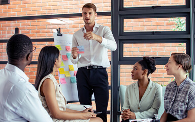 Caucasian smart businessman wearing formal shirt, smiling, presenting project, business plan in front of colleagues, brainstorming, putting post it on whiteboard in meeting room at modern office.