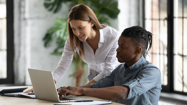 Young Caucasian Businesswoman Help African American Male Intern With Computer Project In Office, Diverse Multiracial Colleagues Brainstorm Work Together On Laptop, Teamwork, Collaboration Concept