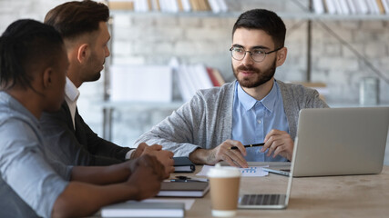 Multiracial young businesspeople sit at desk in office brainstorm at meeting together, diverse multiethnic colleagues coworkers talk discuss business ideas negotiate at briefing, cooperation concept