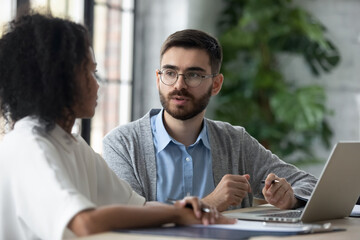 Multiethnic businesspeople sit at desk work on laptop brainstorm discuss business ideas together, multiracial colleagues talk cooperate using computer, negotiate at office meeting, teamwork concept