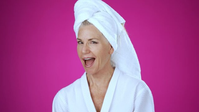 Mature woman in bath robe and towel on head out of shower shocked amazed say wow with winner gesture put hands up senior woman 50s 60s, posing isolated on pink background studio.